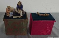 Ceramic figurine group and carved boat displayed on separate velvet-topped brocade boxes
