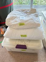 Three stacked bedding items: beige mattress pad, white duvet, cream sheet in clear bin