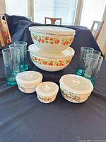 All items grouped: two metal strawberry-pattern mixing bowls with lids, three floral lidded glass bowls, six green tumblers