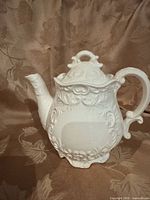 Front view of white embossed ceramic teapot showing spout, handle, lid, and relief design