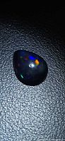 Front view of pear cabochon black opal on textured surface, showing multicolored flashes