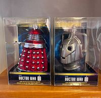 Front view of boxed Doctor Who glass ornaments: red Dalek and silver Cyberman head
