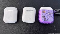 Three Apple AirPods charging cases, one with purple Disney Genie cover