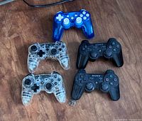 Five controllers laid out: two black Sony DualShock 3 and three PDP Afterglow (1 blue, 2 clear)