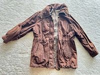 Front view of brown cotton canvas bomber jacket laid flat, showing zipper, pockets, collar, cuffs, and drawstring hem