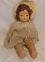 Full view of antique composition baby doll seated, showing dress, bonnet, limbs and overall condition