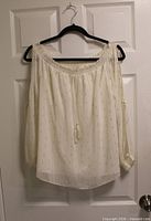 Front view of white Guess off-shoulder blouse with gold dash pattern hanging on hanger