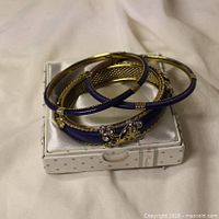 Three gold-tone bangles with enamel and rhinestones stacked on small box