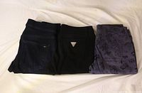 Three folded pairs of jeans: dark wash, black with Guess logo, purple graphic print