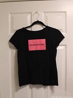 Front view of black Calvin Klein Jeans T-shirt hanging on hanger showing pink logo graphic