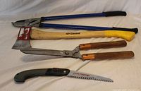 All four garden trimming tools laid out: loppers, axe, hedge shears, folding saw