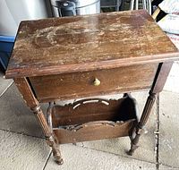 Full view of wooden side table showing drawer, top surface wear, carved magazine rack