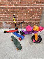 Group shot of trike, two scooters, skateboard against brick wall