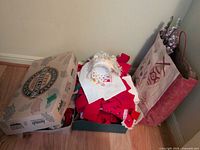 Two boxes and a gift bag holding assorted Christmas bows, stuffed angel, ornaments and floral picks