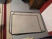 Stacked view of three beige rugs with black trim on floor