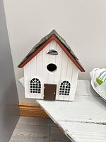 Front view of white wooden birdhouse with brown asphalt shingle roof, arched windows and entry hole