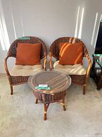 Full view of two rattan chairs with cushions, pillows, and matching round side table