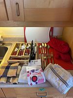 Full lot on countertop including red-handle flatware in wood tray, gold-tone Rogers flatware, cutting board, oven mitts, towels, utensil caddy