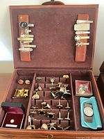 Overall view of open leather case showing tie clips on lid straps, rows of cufflinks, money clip, boxed pins and stud.