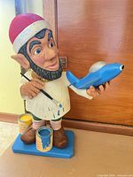 Full view of carved wooden elf statue on base with paint cans and blue airplane