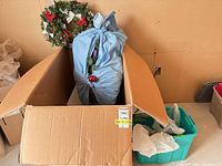 Cardboard box containing bagged artificial greenery, wreath and storage tote