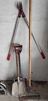 Group shot of garden rake, loppers and two scoop shovels leaning against wall