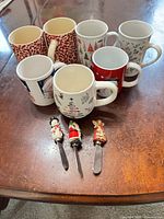 Seven assorted Christmas mugs and three cheese spreaders displayed on tabletop