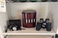 Shelf view showing Nikon Coolpix L27 camera with pouch and cable, wooden photo storage box, and black binoculars