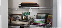 Closet shelf with framed prints, stack of empty frames, puzzle books, cookbooks and albums