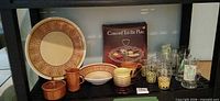 Overall view of lot on shelf showing tid-bit plate box, dinnerware, mugs and glasses