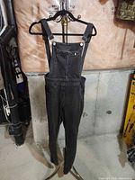 Front view of black denim skinny overalls hanging on stand