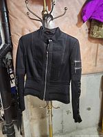 Front view of black jacket on hanger showing zipper, stripe stitching, sleeve lace-up and zip pocket details