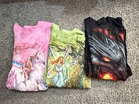Folded pink unicorn, green fairy unicorn, black dragon T-shirts side by side