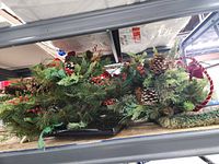 Two of the three wreaths stacked on shelf showing evergreen foliage, pine cones, berries, bows