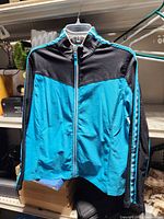 Front view of teal and black lightweight jacket on hanger showing full zipper, pockets, sleeve stripes