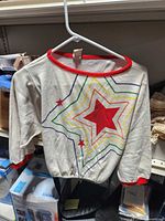 Front view of cream long-sleeve top on hanger showing starburst graphic, red neckline and cuffs