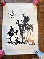 Full view of unframed Picasso Don Quixote poster anchored by cans on wooden floor