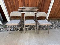 Three padded folding chairs in front of folded table and additional stacked chairs