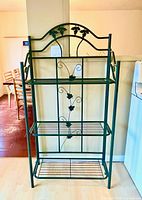 Front view of dark green metal baker’s rack showing three wire shelves and decorative leaf motif