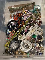 Top view of clear bin filled with assorted costume jewelry showing necklaces, bracelets, earrings, beads and chains