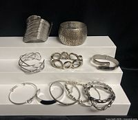 All twelve bracelets and cuffs arranged on tiered white display against black background