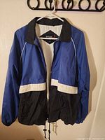 Front view of blue, black and white Sport-Tek windbreaker on hanger