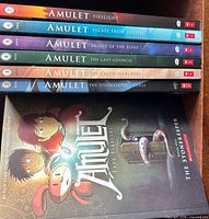 All seven Amulet volumes visible, spines showing numbers 2-7 and front cover of Volume 1