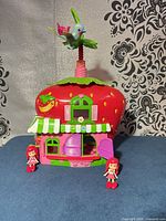 Front view of strawberry playhouse with rooftop hummingbird and two dolls seated outside