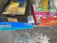 Stack of puzzle boxes showing Van Gogh, Kandinsky, Japanese Tea Garden, Sunflower Field and coasters on floor