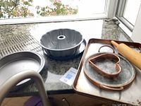 Group of baking items on countertop including Bundt pan, round cake pan, cooling rack, rolling pin, baking tray with heart cookie cutter and copper tray
