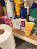 Shelf with assorted cleaning bottles, aerosol cans, trash bag box, paper towel roll and yellow tube