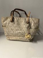 Front view of beige Michael Kors monogram tote bag showing handles, charm and side pockets