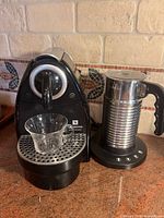 Nespresso Essenza C100 machine with drip tray, clear glass cup in position, Aeroccino 4 frother on adjacent base