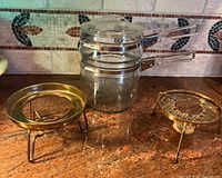 Glass double boiler with lid and two brass tripod stands displayed on countertop